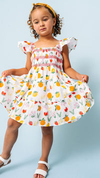 A young girl with curly brown hair wearing a white tiered dress with a colorful fruit print and white sandals, posing against a light blue background.