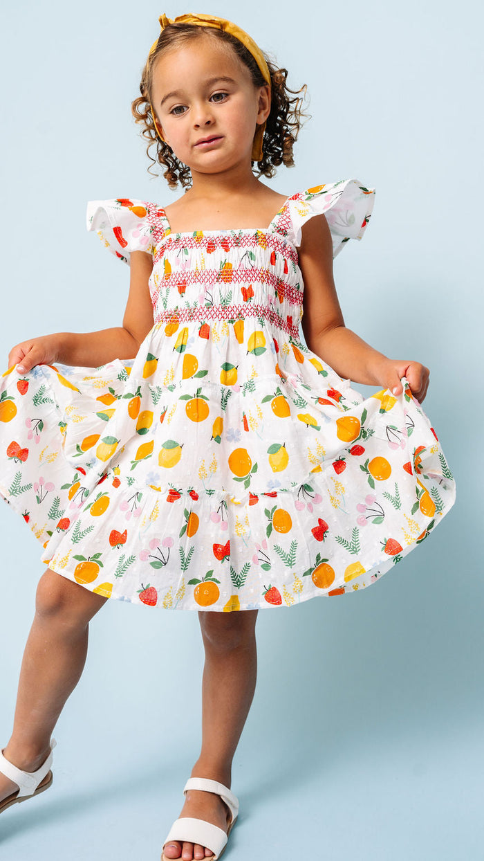 A young girl with curly brown hair wearing a white tiered dress with a colorful fruit print and white sandals, posing against a light blue background.