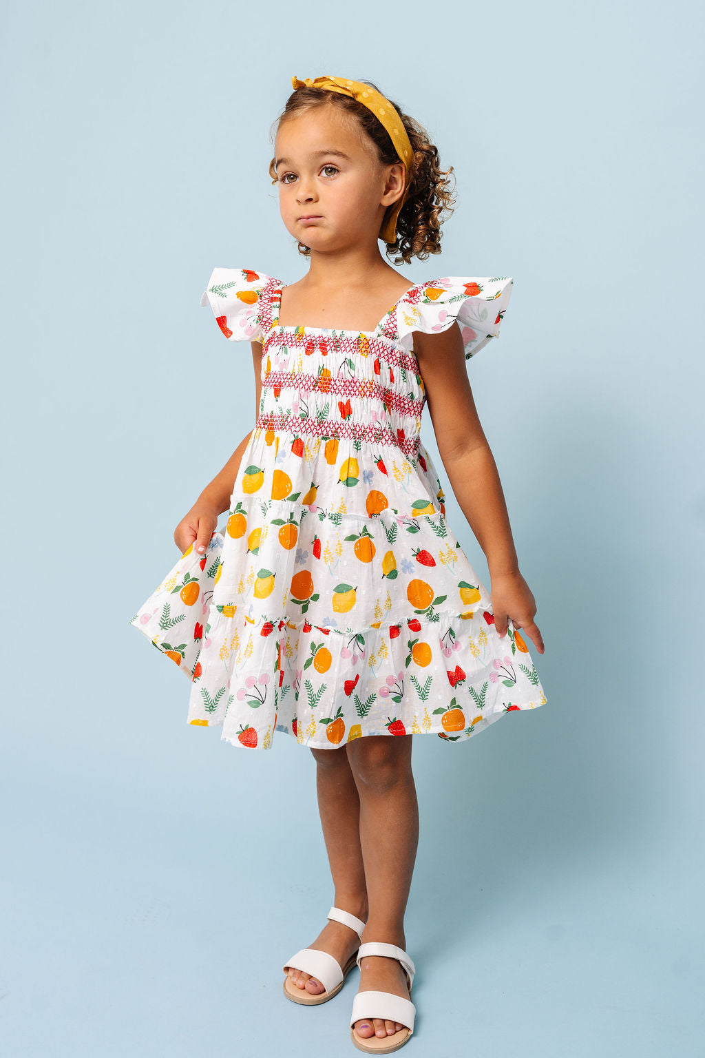 A young girl wearing a white smocked dress with a colorful fruit print, styled with a yellow headband and white sandals, standing against a solid light blue background.