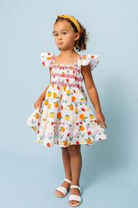 A young girl wearing a white smocked dress with a colorful fruit print, styled with a yellow headband and white sandals, standing against a solid light blue background.